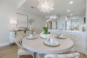 A dining area with a round table, white chairs, a chandelier, and a kitchen in the background.