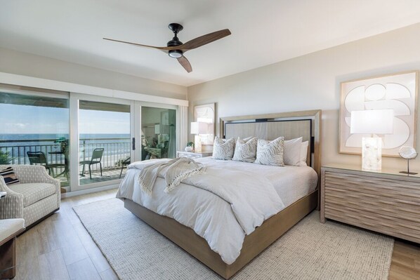 A bedroom with a large bed, a ceiling fan, a nightstand, a lamp, and a view of the ocean through a sliding glass door.