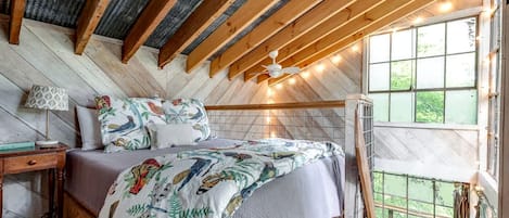 Une chambre avec un plafond en bois, un lit avec une literie à motifs, une table de chevet, une lampe et une fenêtre donnant sur un paysage.