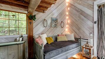 Une chambre à coucher rustique avec un plafond en pente, un lit recouvert de plusieurs coussins, une petite table avec un livre et une fenêtre donnant sur les arbres.