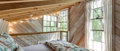 Ein Schlafzimmer mit schräg verlaufender Decke, Holzbalken, einem Bett mit floralem Bettzeug und einem Fenster mit Vorhängen.