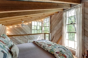 Un dormitorio tipo loft con techo inclinado, cama con ropa de cama floral, ventilador de techo y ventanas grandes con cortinas.