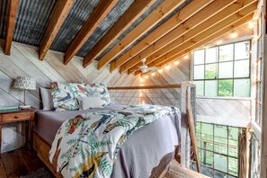 A bedroom with a wooden ceiling, a bed with patterned bedding, a nightstand, a lamp, and a window with a view. - Secluded Tree House in the Forest near the Appalachian Mountains, Georgia (Flintstone)