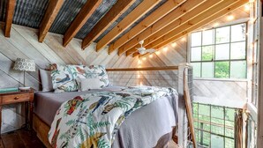 A bedroom with a wooden ceiling, a bed with patterned bedding, a nightstand, a lamp, and a window with a view. - Secluded Tree House in the Forest near the Appalachian Mountains, Georgia (Flintstone)