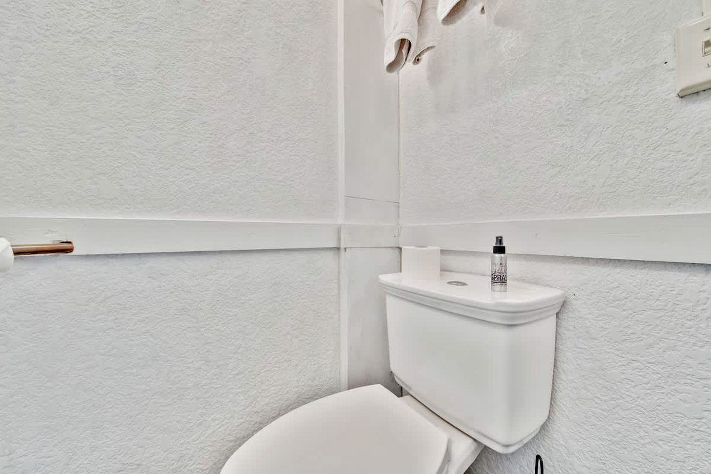 Valkoinen wc-istuin, jonka säiliön päällä on suihkepullo, wc-paperirulla ja metallitangolle ripustettu pyyhe.