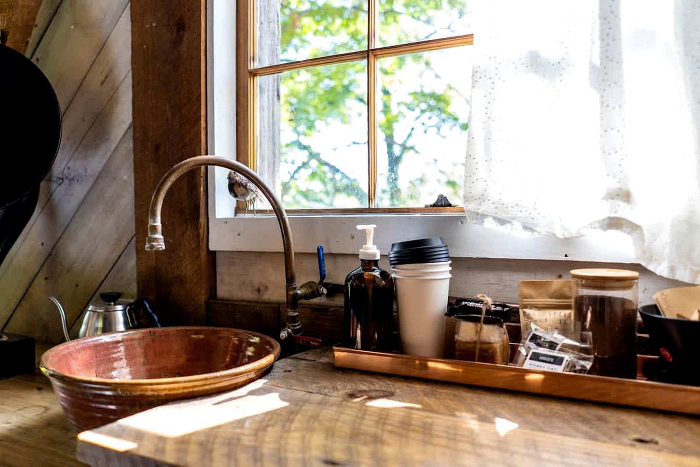 Una cocina rústica con un lavabo de madera, una ventana con vistas a la vegetación y diversos utensilios y recipientes de cocina.