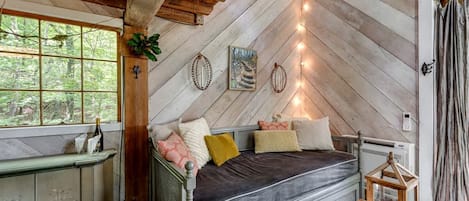 A rustic cabin interior with a loft bed, a small table with a lantern, and a window overlooking greenery.