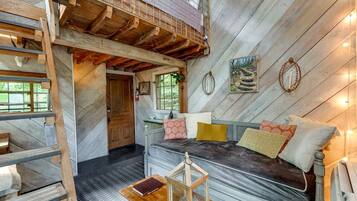 A loft-style bedroom with a loft bed, a wooden ladder, a small table, and a window with a view of greenery.