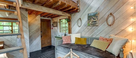 Een slaapkamer in loftstijl met een bedstede, een houten ladder, een tafeltje en een raam met uitzicht op groen.