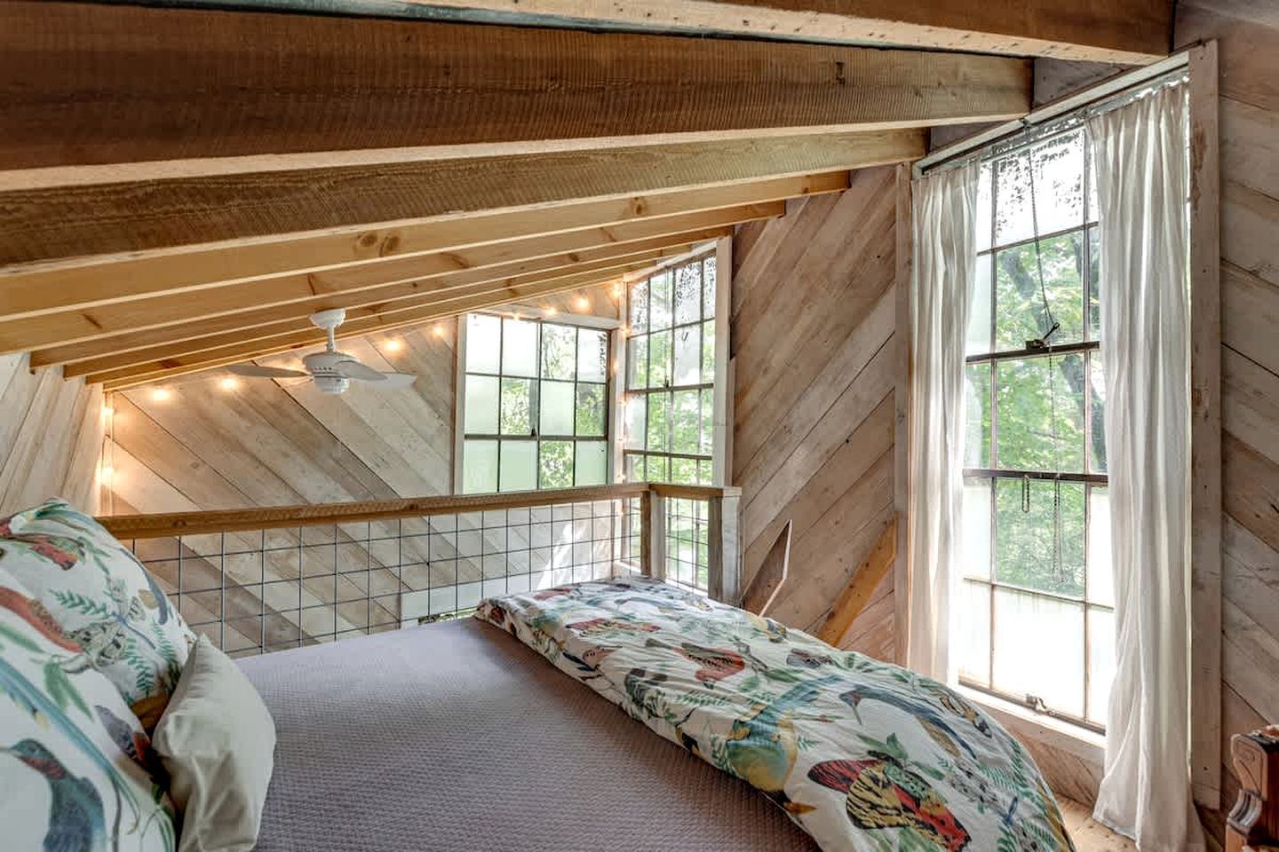 Une chambre située dans un espace de type « loft », avec un plafond en pente, un lit recouvert d’une literie à motifs floraux, un ventilateur de plafond et de grandes fenêtres dotées de rideaux.
