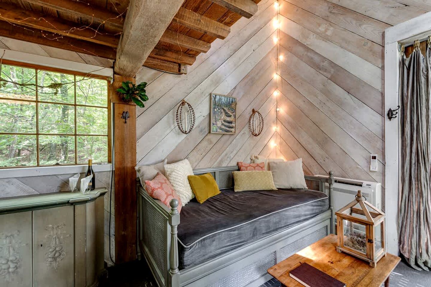 A rustic cabin interior with a loft bed, a small table with a lantern, and a window overlooking greenery.