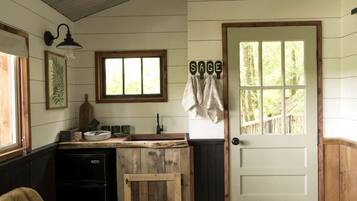 A rustic kitchen with a wooden counter, a sink, and a window. There is a door with a towel hanging on it and a sign that reads 'SAGE'.