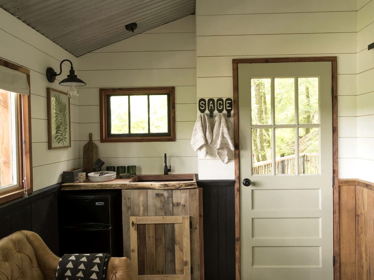 A rustic kitchen with a wooden counter, a sink, and a window. There is a door with a towel hanging on it and a sign that reads 'SAGE'.