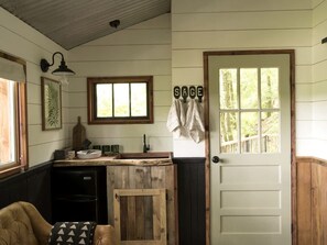 A rustic kitchen with a wooden counter, a sink, and a window. There is a door with a towel hanging on it and a sign that reads 'SAGE'.