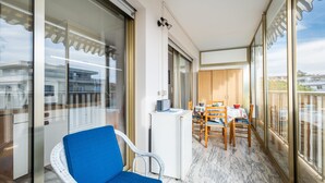 A balcony with a blue chair, a dining table with chairs, a small refrigerator, and a view of buildings through large glass doors.