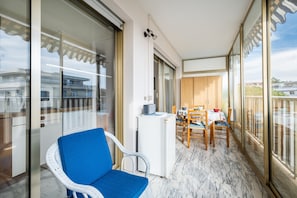 Een balkon met een blauwe stoel, een eettafel met stoelen, een kleine koelkast en uitzicht op gebouwen door grote glazen deuren.