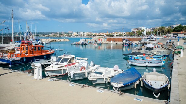 A marina with various boats docked, a clear sky, and buildings in the background.