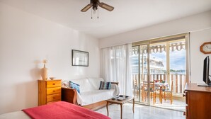 A bedroom with a bed, a dresser, a nightstand, a window with curtains, a balcony with a table and chairs, and a ceiling fan.