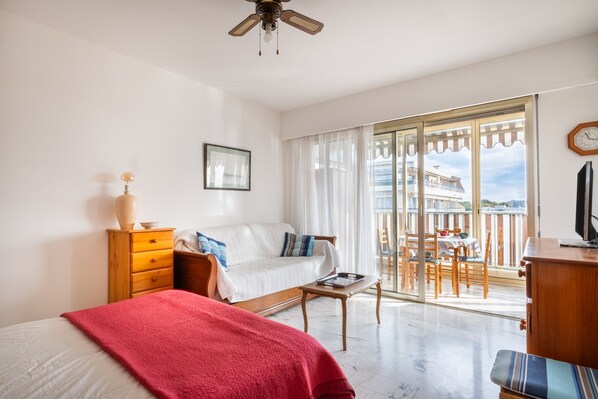 A bedroom with a bed, a dresser, a nightstand, a window with curtains, a balcony with a table and chairs, and a ceiling fan.