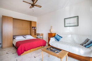 A bedroom with a bed, a sofa, a wooden wardrobe, a ceiling fan, a small table, and a framed picture on the wall.