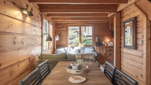 A wooden cabin interior with a dining table, chairs, a sofa, and a window with a view of greenery.