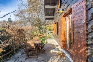 A wooden cabin with a patio table and chairs, a wooden deck, and a window with a light fixture.
