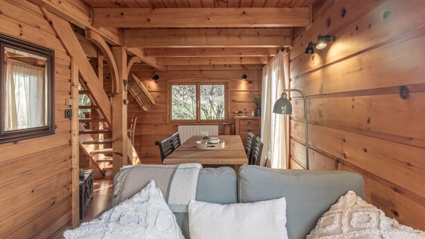 A wooden cabin interior with a dining area, a sofa with cushions, and a staircase.