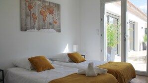 Un lit double avec une literie blanche et moutarde, une carte du monde accrochée au mur et une porte-fenêtre coulissante donnant sur un espace extérieur.