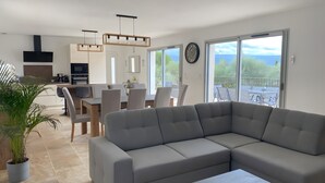 Un salon moderne avec un canapé d’angle, un coin repas avec des chaises, une cuisine équipée d’un téléviseur et de grandes fenêtres offrant une vue dégagée.