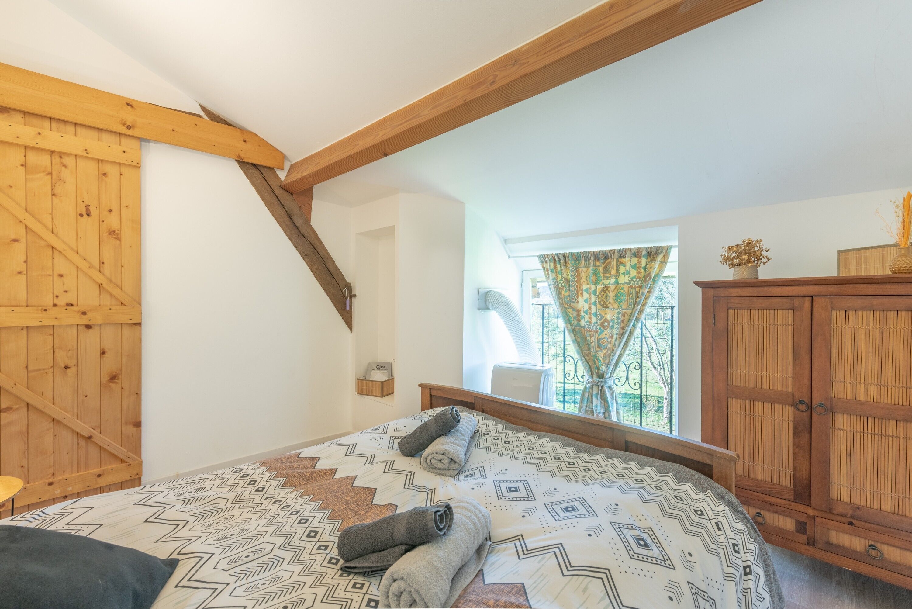 Una camera da letto con soffitto in legno, un letto con biancheria a motivi, una finestra con tende e un armadio in legno.