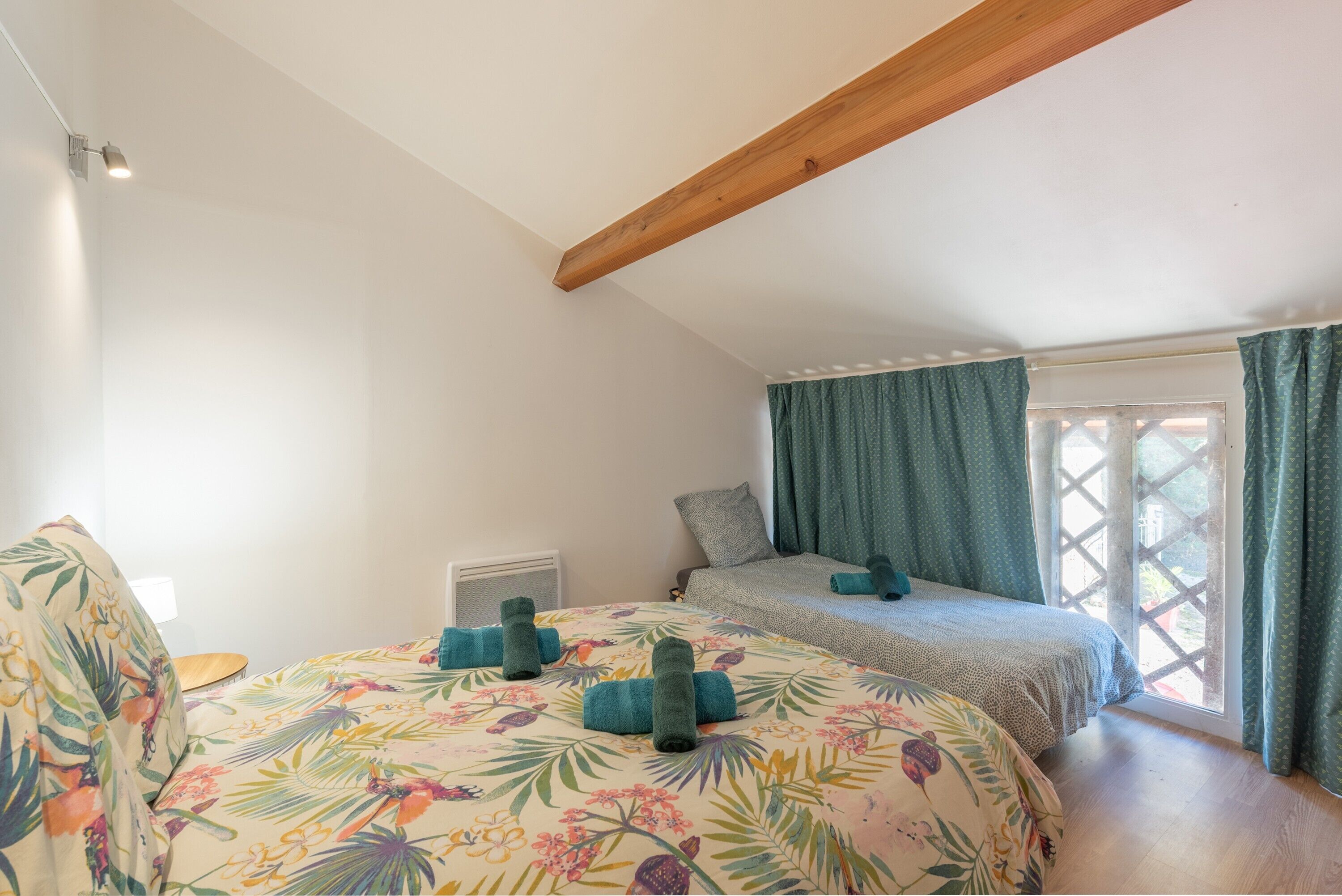 Une chambre avec un plafond en pente, un lit avec une literie à motifs floraux, une fenêtre avec des rideaux et un radiateur.