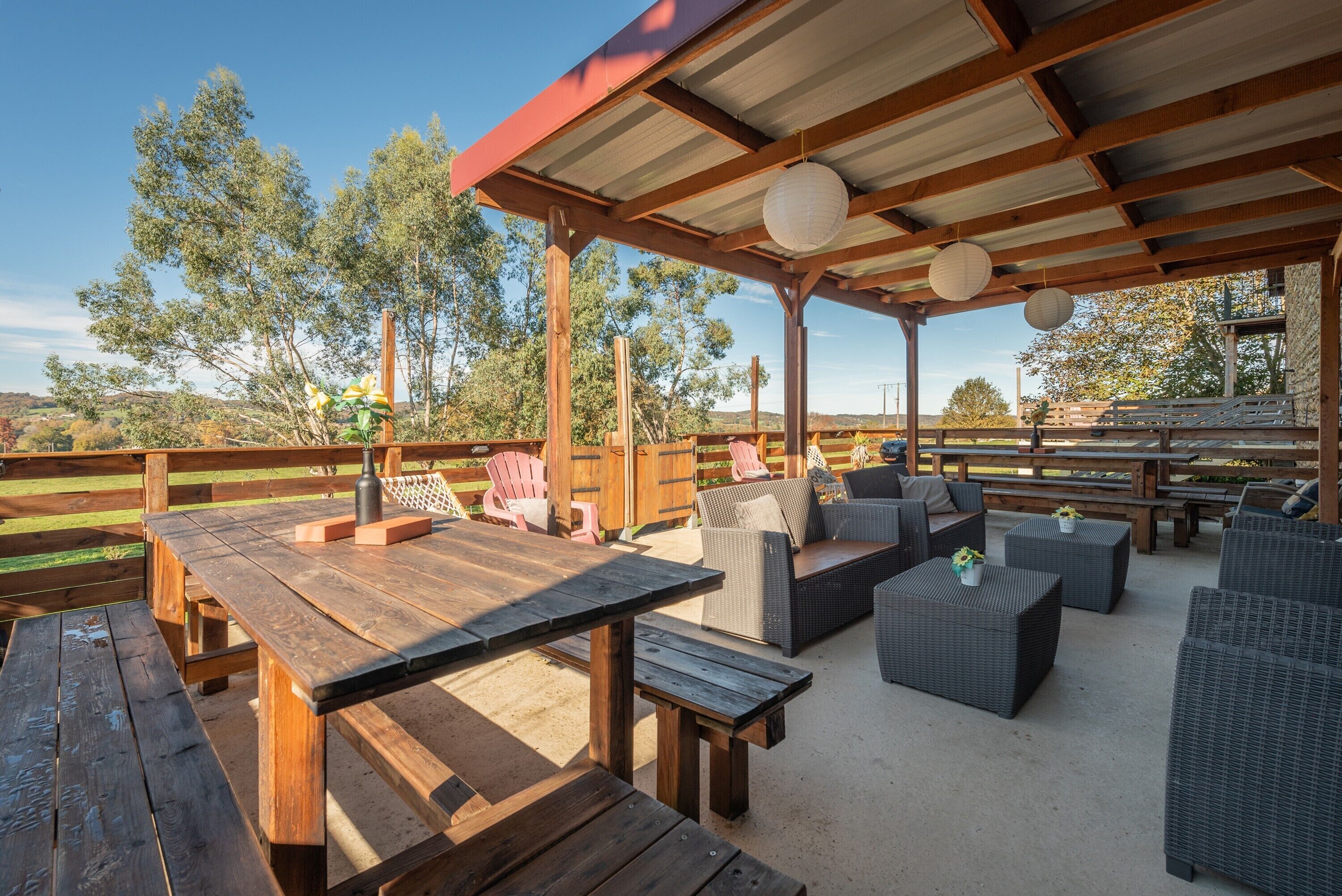 An outdoor patio with wooden tables and wicker furniture, surrounded by a wooden fence and trees.
