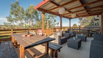 An outdoor patio with wooden tables and wicker furniture, surrounded by a wooden fence and trees.