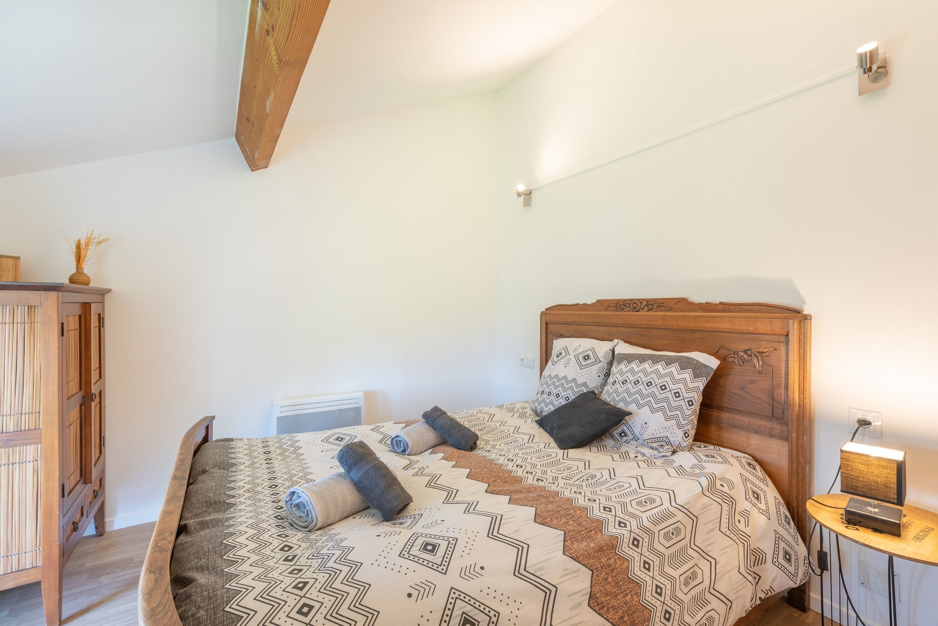 A bedroom with a wooden headboard, patterned bedding, a bedside table with a lamp, and a wooden wardrobe.