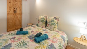 A bedroom with a bed, floral bedspread, two pillows, a wooden wardrobe, a small round table with a lamp, and a wall-mounted light. - Holiday Home “Galets” with Mountain View, Wi-Fi, and Air Conditioning (Peyraube)