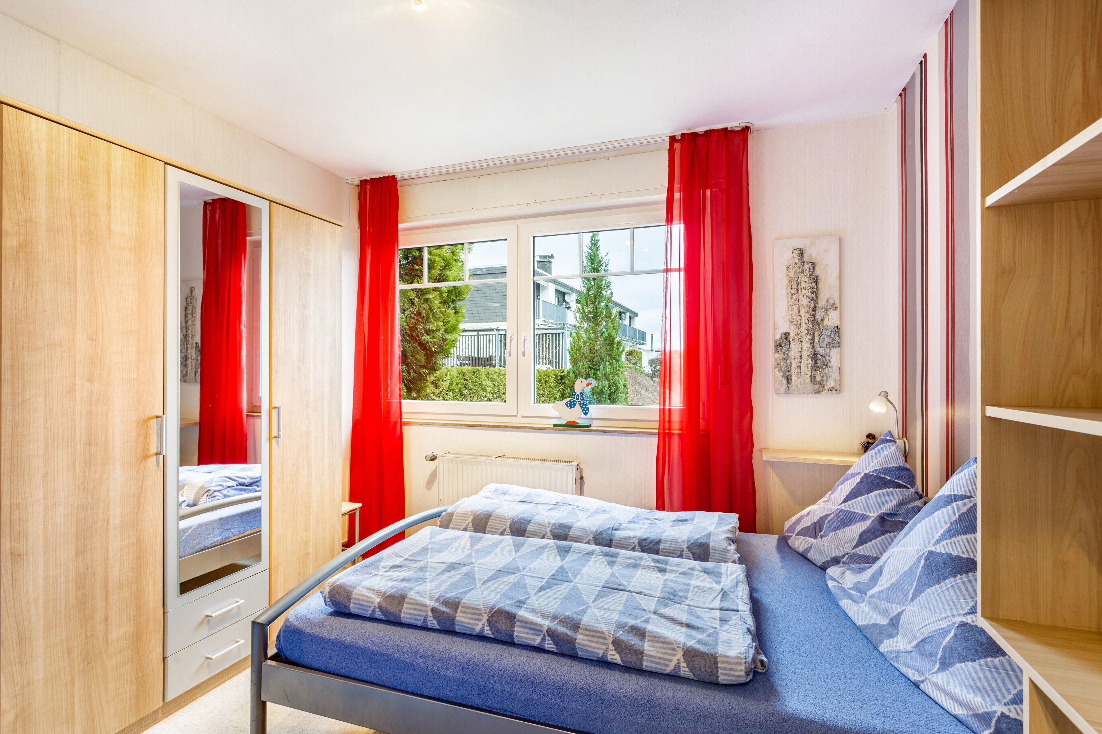 Ein Schlafzimmer mit Bett, Kleiderschrank, einem Fenster mit Vorhängen und Blick auf Gebäude.