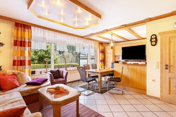 Ein gemütliches Wohnzimmer mit Sofa, Esstisch und Stühlen, einem Fernseher und einem Fenster mit Vorhängen.