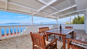 A covered terrace with a table and chairs, a view of the sea, and a white railing.