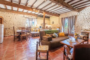 A rustic interior with exposed wooden beams, stone walls, a dining area with chairs and a table, and a living room with a sofa and cushions.