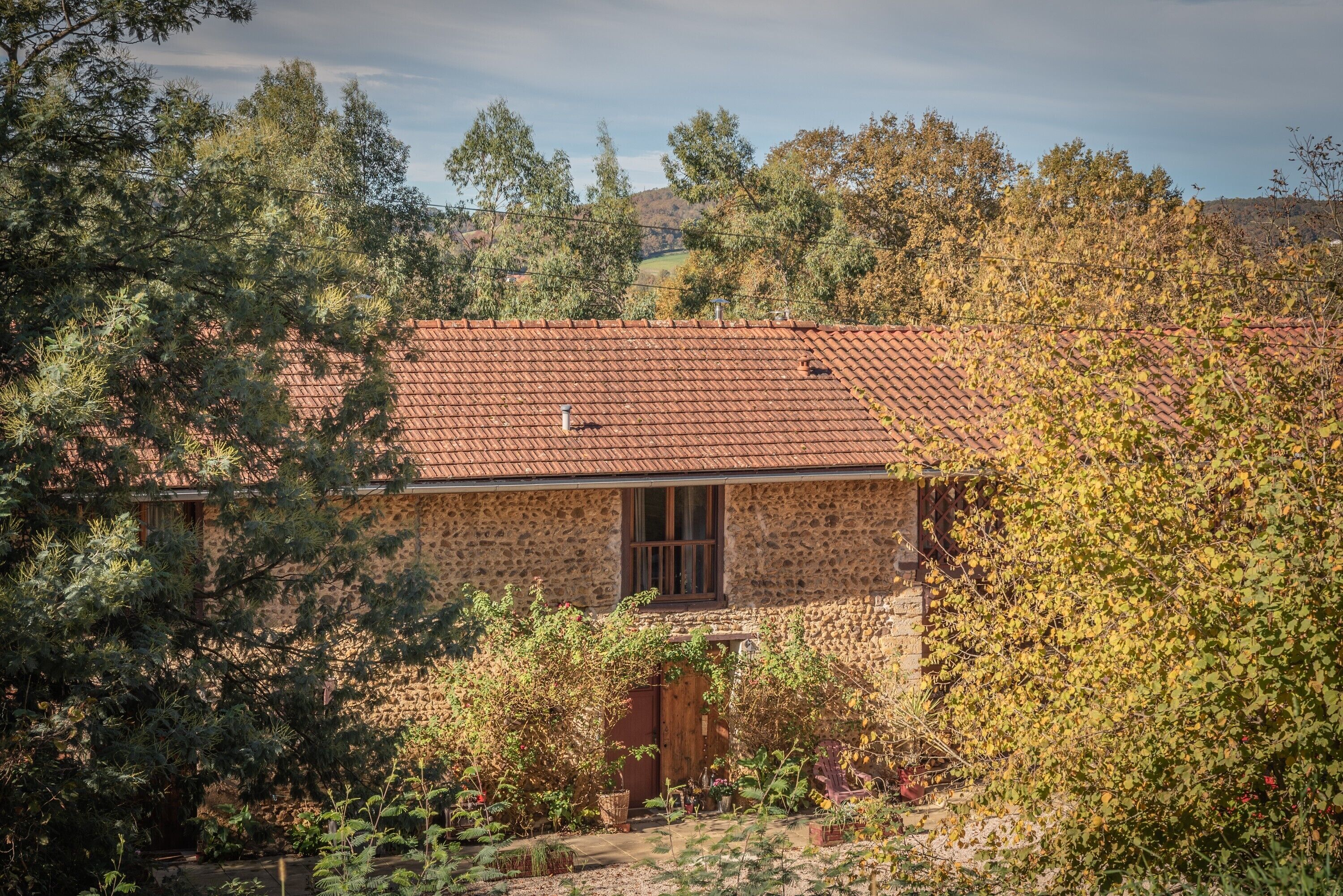Une maison en pierre avec un toit de tuiles, entourée d’arbres et de végétation.