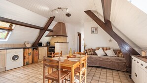 A kitchen with a dining table, chairs, a washing machine, and a sofa in a loft-style room with exposed beams and a tiled floor.