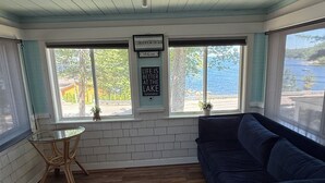 A room with a dark blue sofa, a glass table, and a ceiling fan. - Sebago Lake studio-style condo with enclosed sunroom and sunset views (Standish)