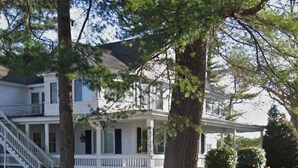 A two-story white house with a covered porch and a staircase leading to the second floor. - Sebago Lake studio-style condo with enclosed sunroom and sunset views (Standish)