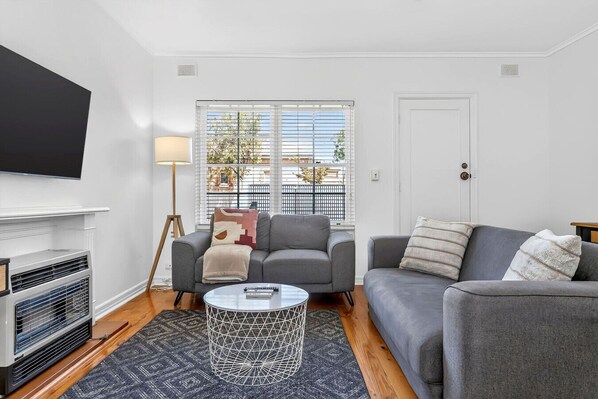A living room with a grey sofa, a coffee table, a TV mounted on the wall, and a window with blinds.