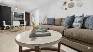 Un salon moderne avec une table basse ronde, un canapé beige agrémenté de coussins bleus et un coin repas avec des chaises blanches.