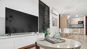 Un salon moderne doté d’un téléviseur à écran plat, d’une table de salle à manger ronde et d’un coin cuisine avec des meubles blancs et un four à micro-ondes.