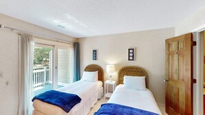 A hotel room with two beds, a balcony, a wooden door, and a small table with a lamp.