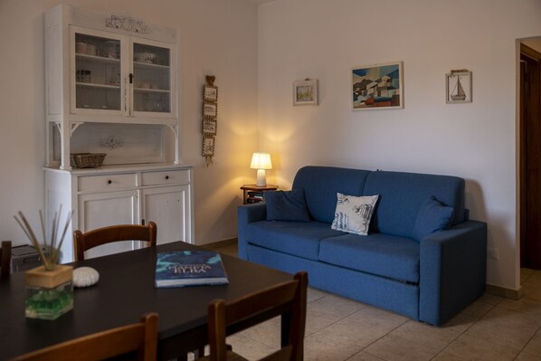 A living room with a blue sofa, a dining table with chairs, a white cabinet with glass doors, and a wall with framed pictures.