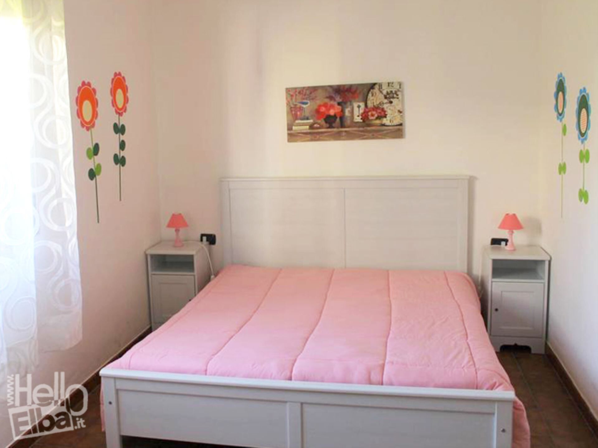 A bedroom with a pink bedspread, white headboard, two bedside tables with lamps, and wall decorations.