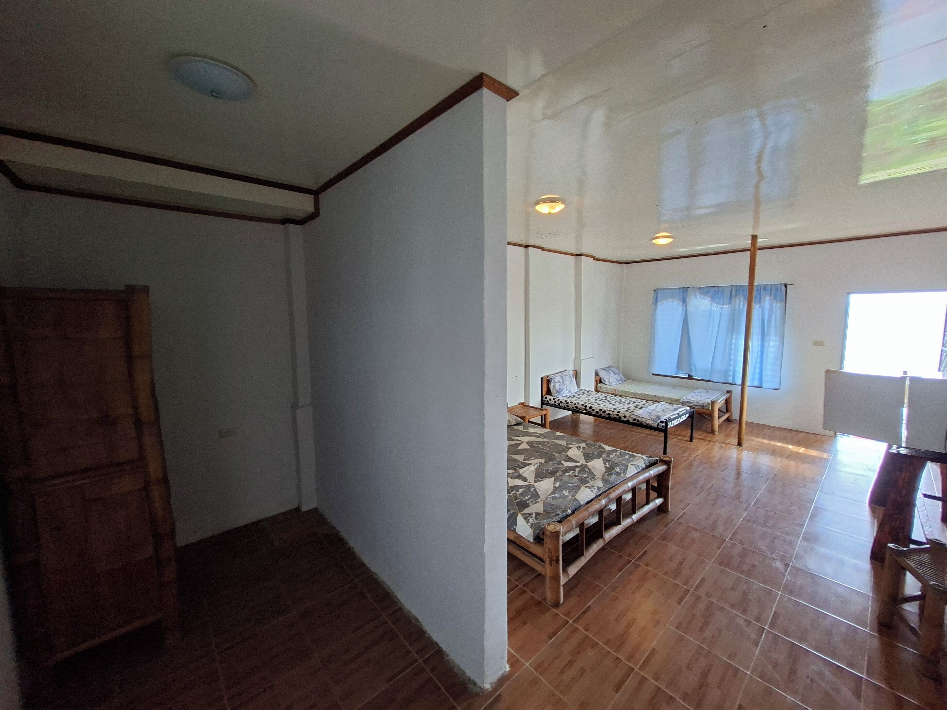 A room with a wooden wardrobe, a bed with a patterned mattress, a bench, and a window with curtains.